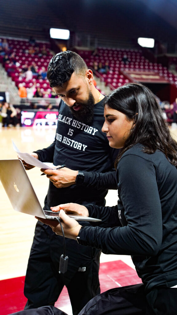 DERRICK SLAYTON, VIDEO COORDINATOR AT BOSTON COLLEGE WBB and staff discuss strategies courtside, with a laptop showc indicating the CatapultPro Video Focus software in foreground.この画像は、ライブ・トゥ・ベンチのワークフローが、ゲームタイムの調整と戦略立案を即座に促進することの本質を捉えている。