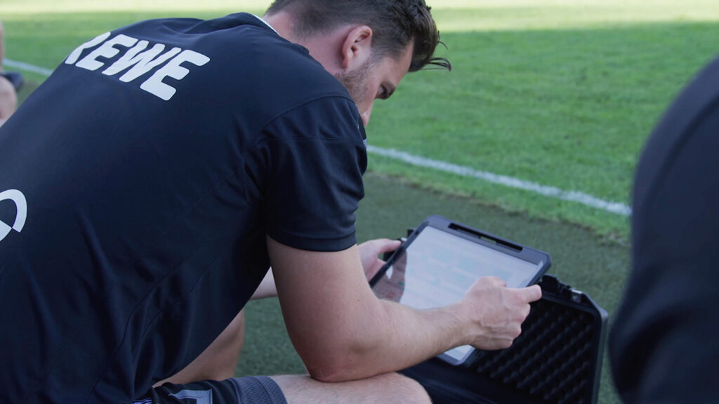Futbolista de negro revisa datos tácticos en una tableta con un entrenador durante una sesión de entrenamiento, con un campo como telón de fondo.