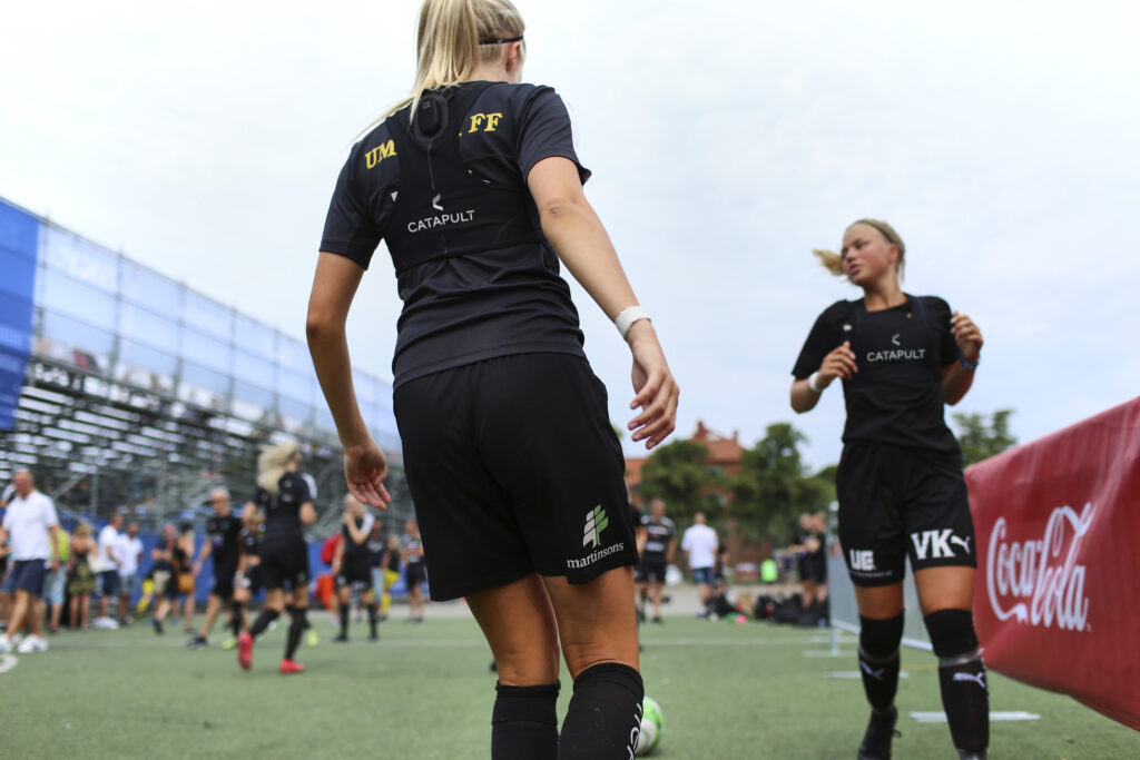 Female football players wearing Catapult performance vests during a pre-match warm-up session, preparing to monitor physical performance and tactical execution.