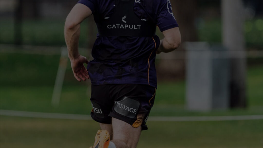 Vista trasera de un jugador de la NRL a media zancada en el campo de entrenamiento, mostrando el dispositivo tecnológico Catapult en su camiseta morada y negra. El escenario es al aire libre y se centra en la forma atlética del jugador y el equipo de entrenamiento de alta tecnología.