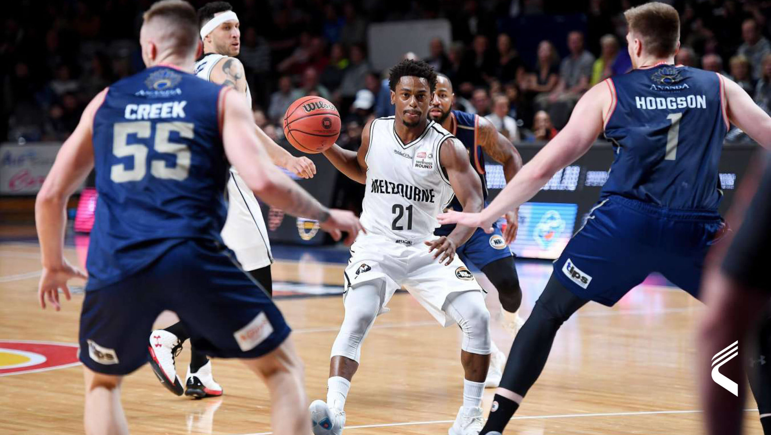 A Melbourne United kosárlabdázója, aki fehér mezben, 21-es számmal ellátott mezben dribbel a labdával, miközben két kék mezes ellenfél játékos szorosan őrzi. A játék intenzív, a játékosok a támadásra és a védekezésre egyaránt összpontosítanak, miközben a pályán mozognak.