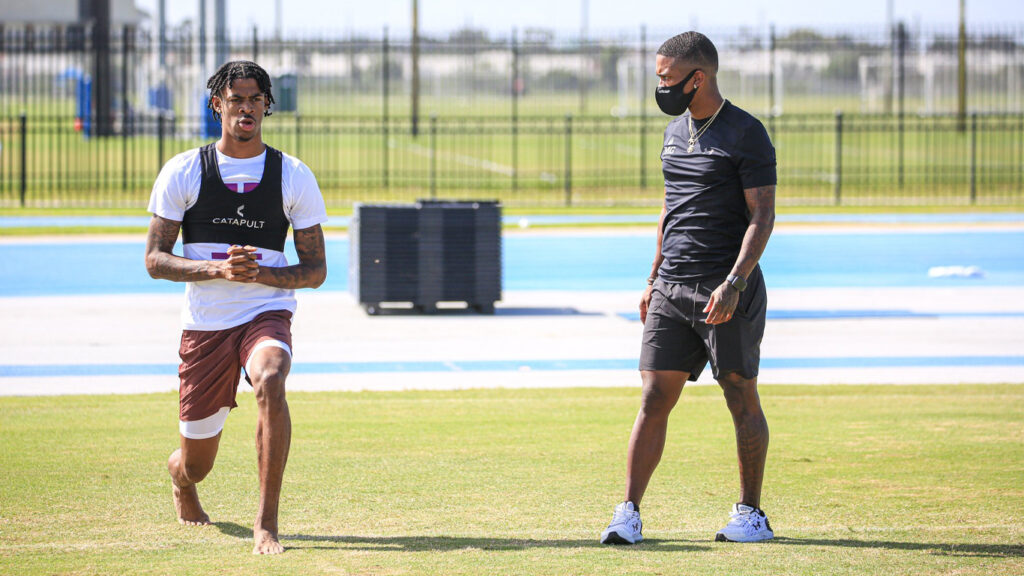 Atleta masculino con un chaleco blanco y morado con la marca "CATAPULT" entrena en un campo soleado, vigilado por un entrenador vestido de negro.