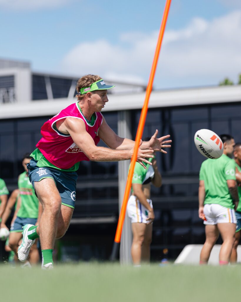 Hráč týmu Canberra Raiders v růžové tréninkové vestě podává ragbyový míč během tréninku, v pozadí jsou spoluhráči a tréninkové vybavení.