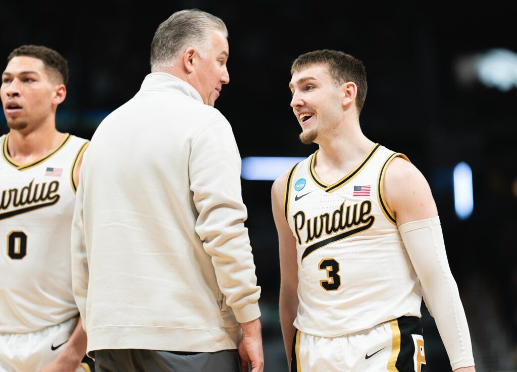 L'allenatore di pallacanestro dei Purdue Boilermakers con una felpa beige parla con un giocatore sorridente che indossa una divisa bianca e oro con il numero 3, a bordo campo.